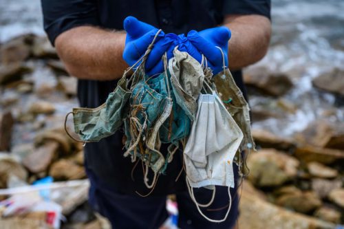 A person wearing bright blue gloves stands on rocks next to a body of water holding dirty individual masks that have become waste.