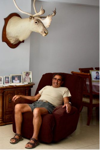 A person in a white shirt, blue jean shorts, and dark sandals sits in a brown reclining chair surrounded by framed children’s photos and a mounted deer head on the wall above.