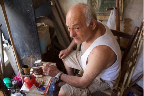 A person in a white sleeveless shirt and beige shorts sits on a wooden chair warming a metal container on a small gas stove.