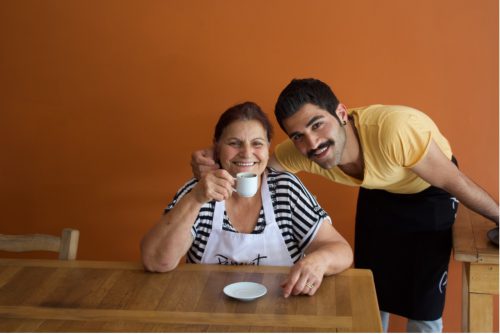 A person wearing a black-and-white striped T-shirt and a white apron holds up a small white teacup. Another person in a yellow T-shirt, blue jeans, and a black apron bends down to them with their arm around the other person’s shoulders.