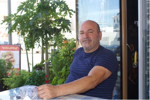 A person with graying facial hair wearing a blue striped shirt sits at a glass-top table in front of short trees and bushes, and a sign with food on it.