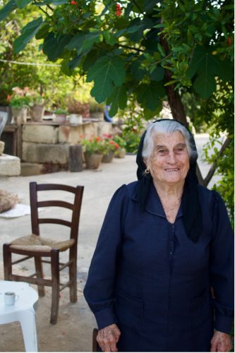 A person with white hair wearing a dark dress and a black head covering stands under a tree in front of a brown chair and plants in stone pots.