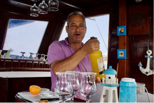 A person wearing a pink shirt holds a large bottle of yellow liquid and stands behind a table with salt-rimmed glasses, half an orange, and other objects on it.