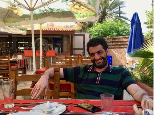 A person wearing a dark blue and green striped shirt sits at a table with a bright red table cloth on a restaurant patio under an open umbrella.