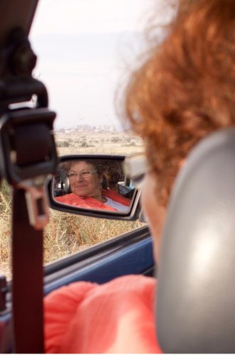 A photograph taken in a car over the shoulder of a person wearing an orange shirt and glasses shows the person's face in the sideview mirror.