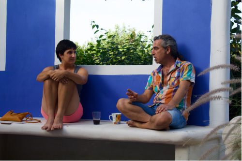 A person wearing a blue tank top and pink shorts sits with their knees to their chest and talks to another person in a multicolored shirt and blue jean shorts as they lean on a blue and white wall.