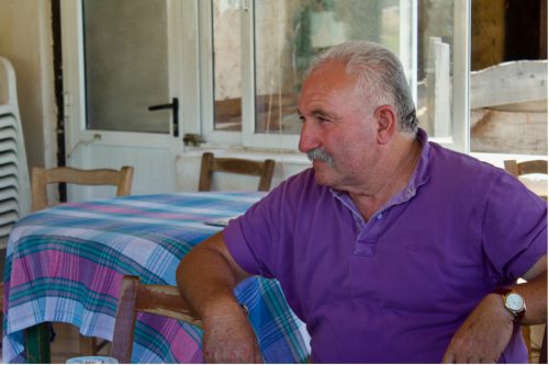 A person wearing a purple shirt and a brown watch rests their arm on a wooden chair in front of a table with a purple and blue plaid tablecloth on it.