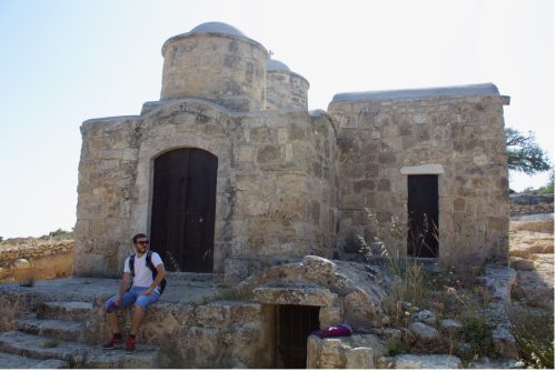 A person wearing a white T-shirt, blue jean shorts, and sunglasses sits on the steps of an old stone building with large brown doors.