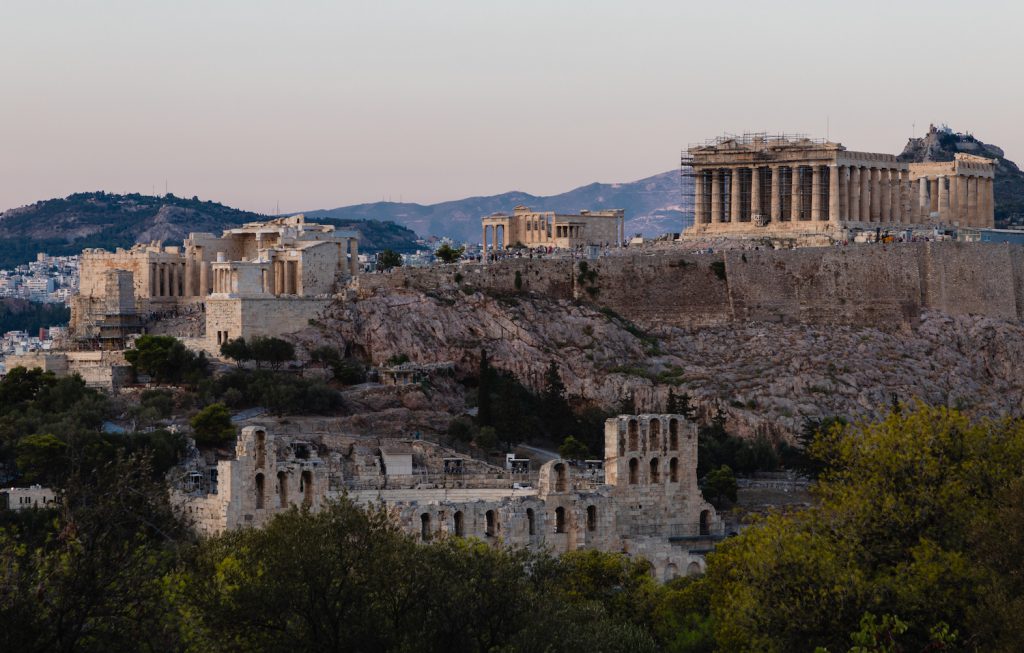A group of white stone buildings, with the Acropolis complex at the top, sit on a hill surrounded by trees at sunset.