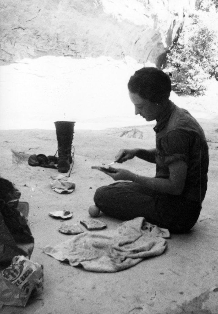 A black-and-white image of a pioneering female archaeologist shows her wearing a shirt with rolled-up sleeves and sitting in the shade spreading food on a slice of bread.