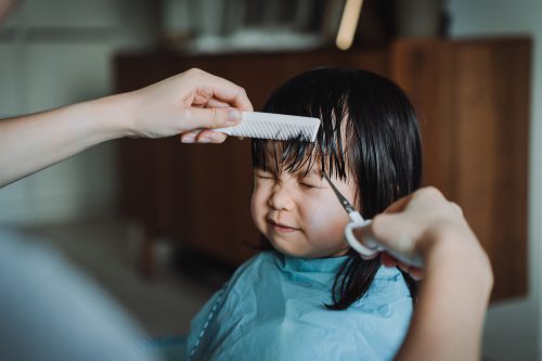 A young child with black hair and a blue cloth over their body scrunches their face as someone uses a white comb and white scissors to cut their hair.