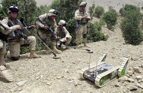 Four soldiers in military gear kneel and stand as they watch a small robot in a sandy clearing surrounded by brush.
