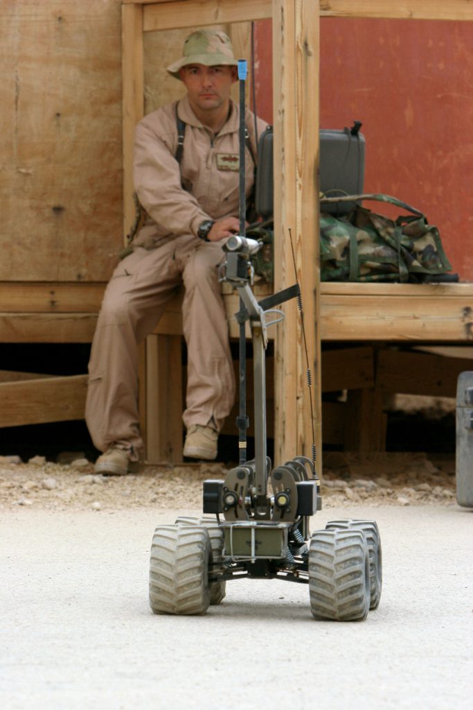 A person dressed in tan fatigues sits on a bench behind a robotic device equipped with heavy-duty wheels and a long metal pole with a camera attached.