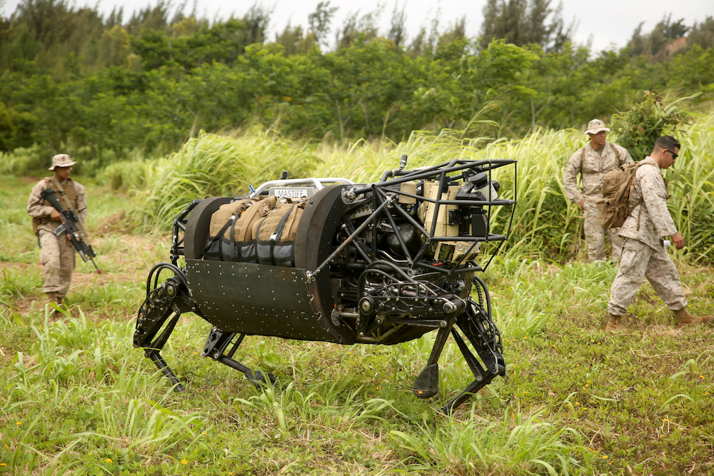 A large, dark metal robot with four legs and brown bags secured inside it walks alongside three soldiers in tan uniforms surrounded by grasses and trees.