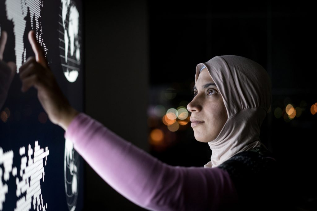 A person wearing a pink shirt and a light-pink hijab stands in a dark space with their face illuminated while pointing to images of data on a large screen.