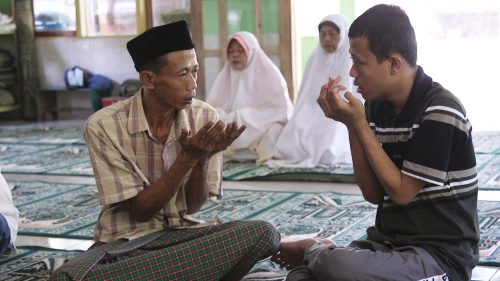An older man sits with a younger man inside on teal patterned rugs, both with their hands cupped before them in prayer.