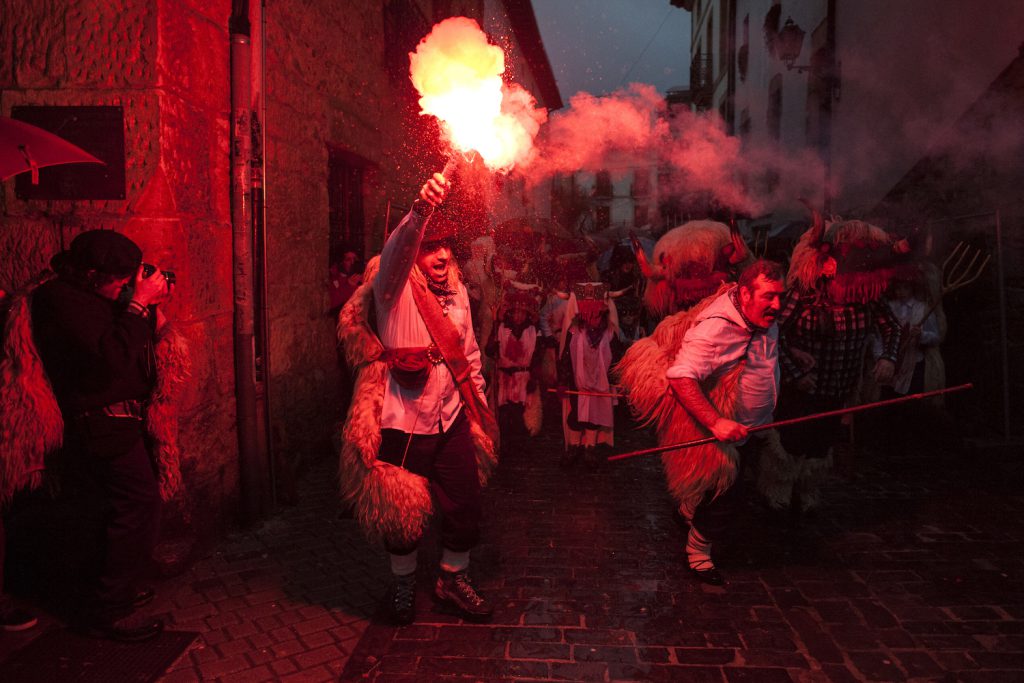 A group of people with white tunics and animal pelts on walk up a narrow street. One person holds a flare that gives off a red glow. A person wearing dark clothing photographs the event.
