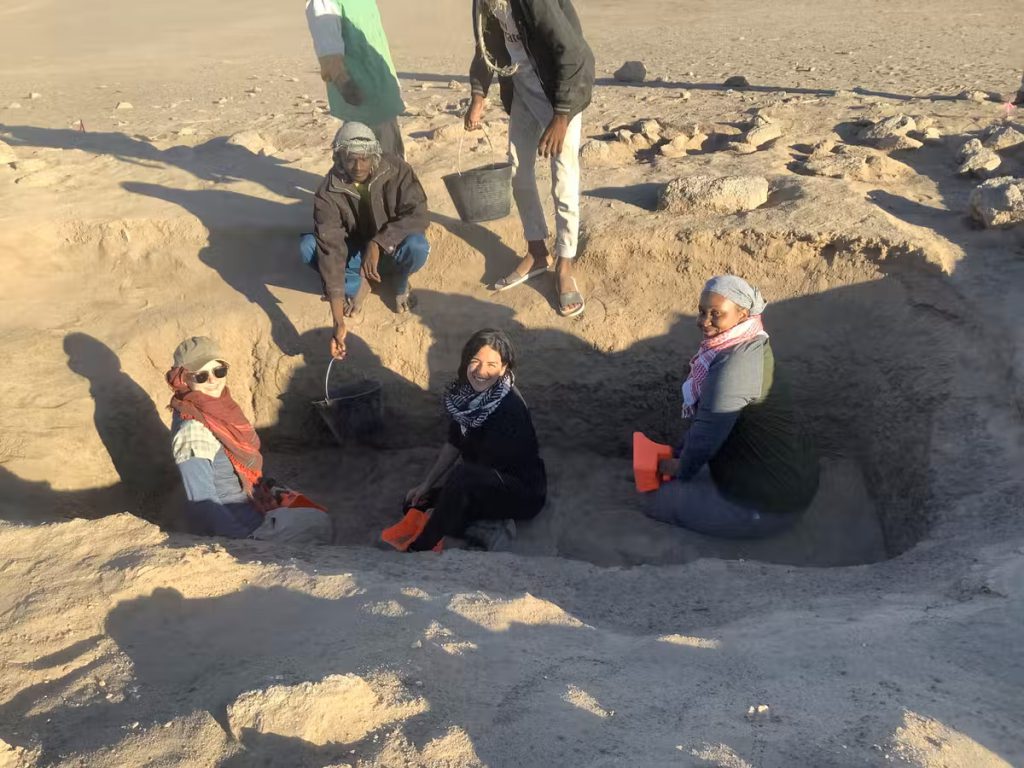 Three people sit in an excavation pit, working to uncover ancient Nubia, and look back at the camera while three others stand on the sand and approach the platform.