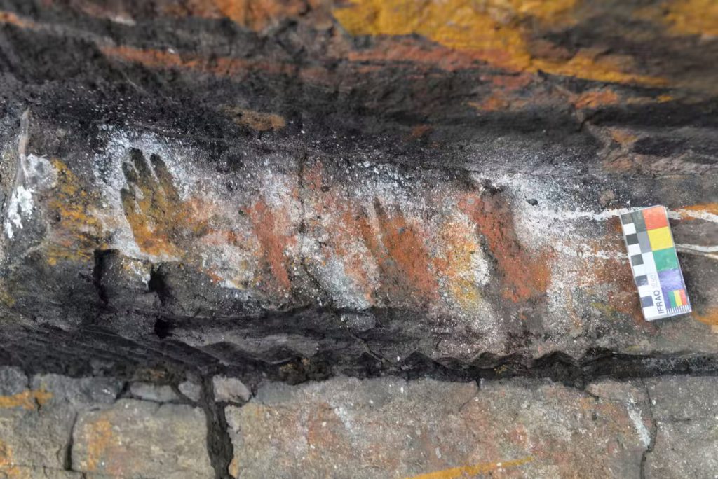 Four hand shapes are shown on black and brown stone outlined with white paint next to a black, white, red, yellow, purple, and green measuring stick.