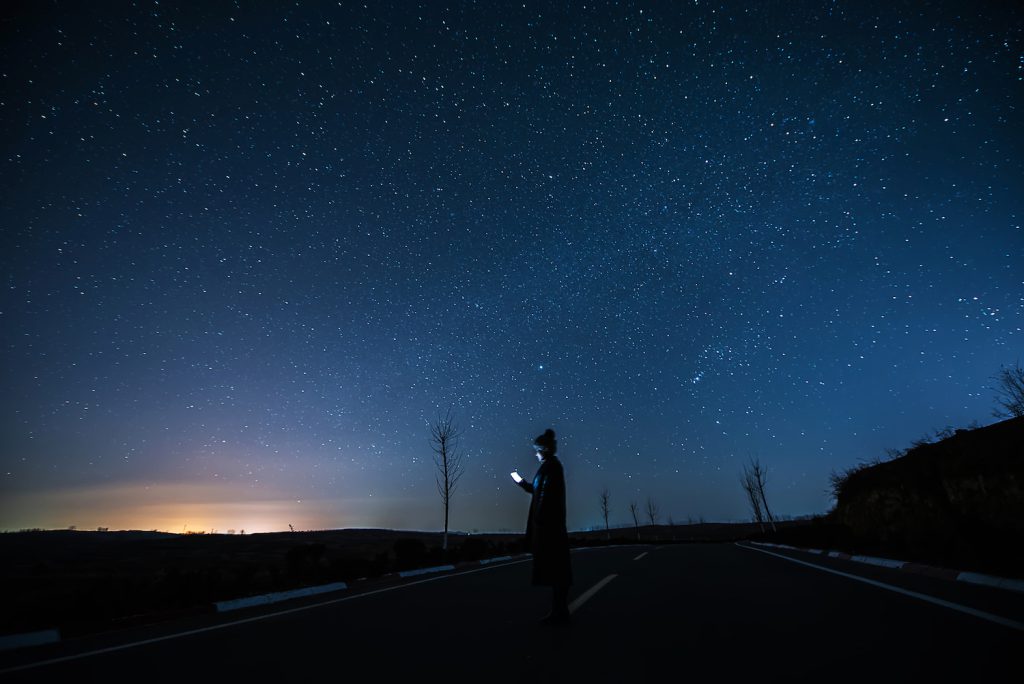 A person stands outside under a dark-blue, starry sky and looks at the bright screen of their cellphone. A yellow light shines in the distance.