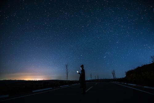 Una persona se para afuera bajo un cielo estrellado azul oscuro y mira la pantalla brillante de su teléfono celular. Una luz amarilla brilla en la distancia.