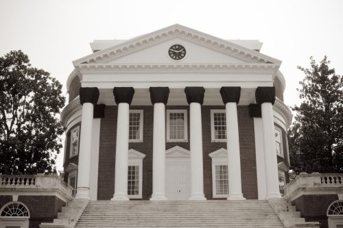 A dark building is featured with white trimmed windows, a white door, and six white columns underneath a dark clock face.