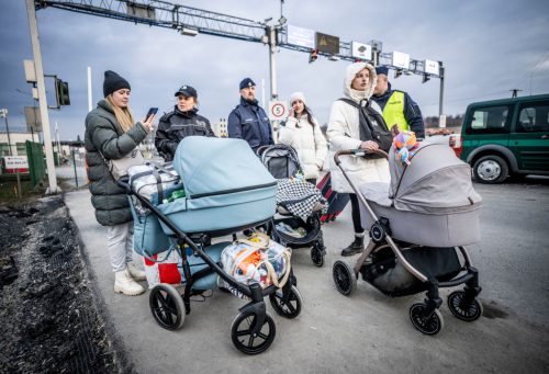 Several women wearing heavy coats push baby carriages on a path at a border checkpoint, with people dressed in uniforms walking behind and next to them.