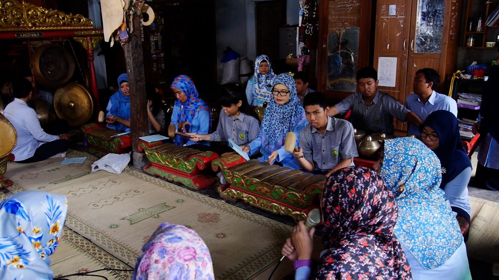 A group of people—mostly women in colorful headscarves and a few men wearing button down shirts—sit in a circle on mats on the floor, watching students play instruments.