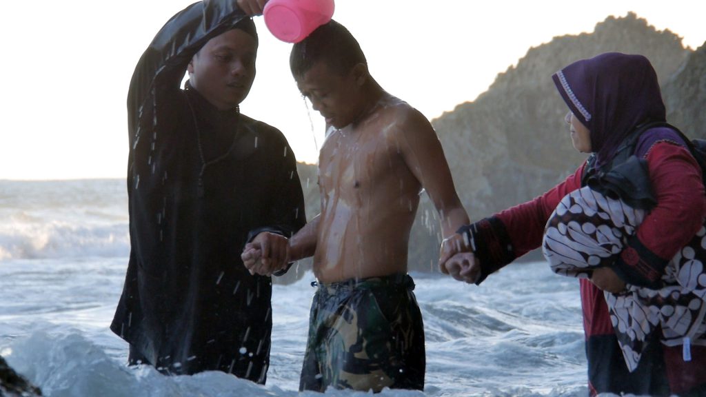 Three people stand nearly waist-deep in the ocean; the person on the left pours water over the shirtless young man in the center, who holds hands with a woman standing on the other side.
