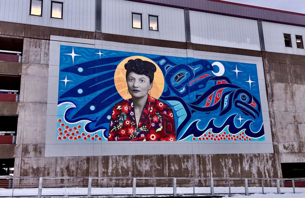 A mural shows a woman wearing a colorful shirt with a yellow orb around her head surrounded by blue waves, the blue outline of a bird, and the dark-blue figure of the head of a salmon against stars and the moon.