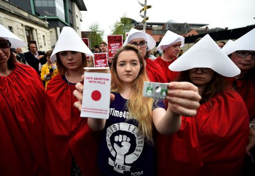A person with blonde hair and a black-and-white shirt with a fist on it stands among a group of people who wear red robes and white triangular hoods. She holds up a silver blister package of pills and a white box with red writing that says, “Safe abortion with pills.”