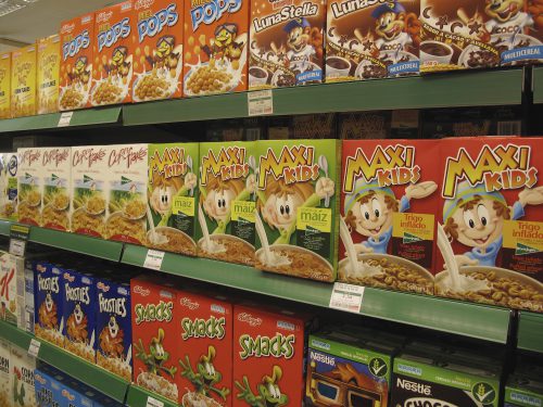 Grocery store shelves are shown packed with colorful cereal boxes adorned with cartoon animals and children, as well as labels in Spanish and English.