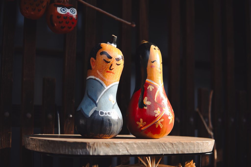 Two yellow gourds painted to look like a frowning man with black hair wearing a blue top and a smiling woman with long black hair and a red and white dress sit on a ledge.