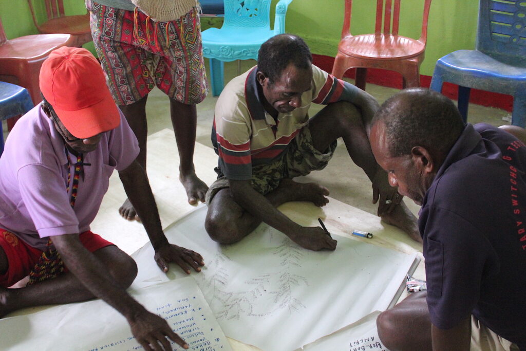 Three people sitting on the floor point to and draw on large white pieces of paper. Another person stands behind them.