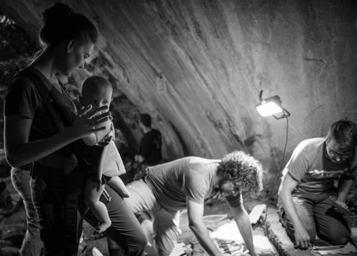 Una imagen en blanco y negro muestra a una mujer que lleva a un bebé en un arnés frontal mirando a dos personas que examinan objetos en el suelo de una cueva bajo una luz de pie.