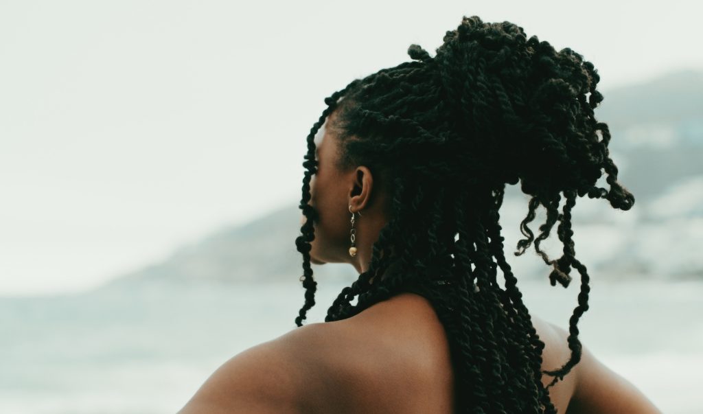 A person with many small, long, black braids tied up and flowing down their back looks out onto the water.