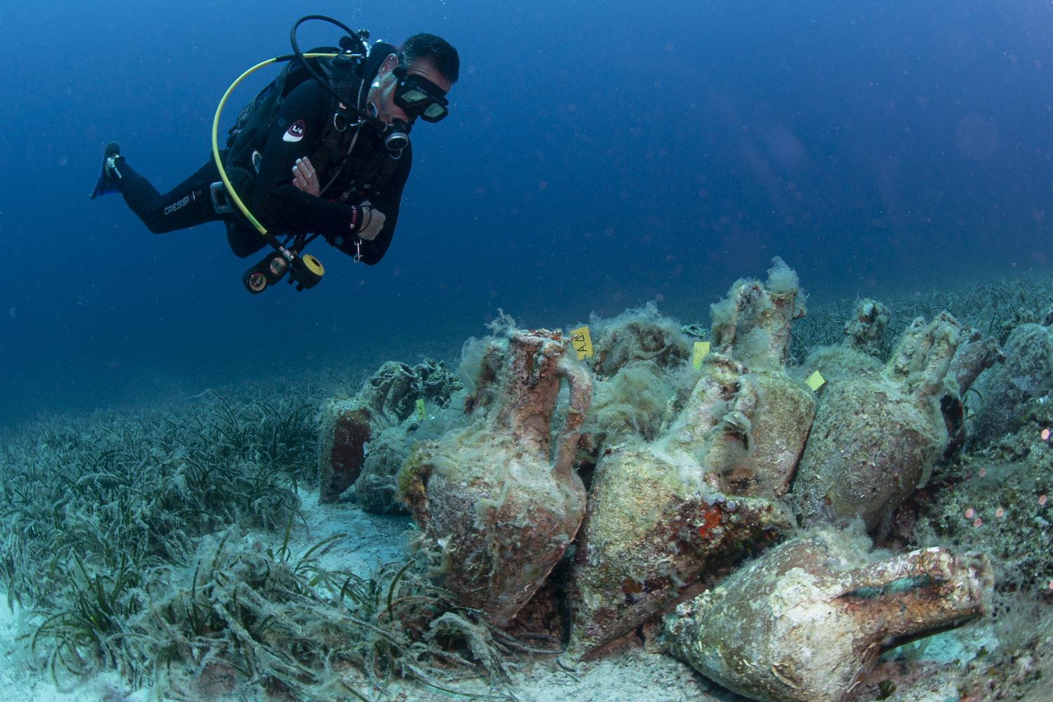 A person in a scuba suit hovers underwater above a cluster of large vases covered with algae and moss resting on the sea floor.