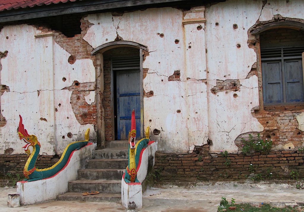 A white temple wall with partially exposed brick is covered with holes. Two red, green, and yellow dragons perch on either side of stairs leading to a blue door.