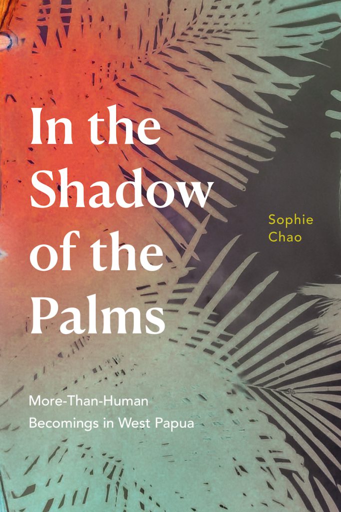 A book cover shows green and red palm fronds resting on top of a dark gray background, with the title In the Shadow of the Palms in large white font. The subtitle in white reads “More-Than-Human Becomings in West Papua,” and the author’s name, Sophie Chao, is in yellow font.