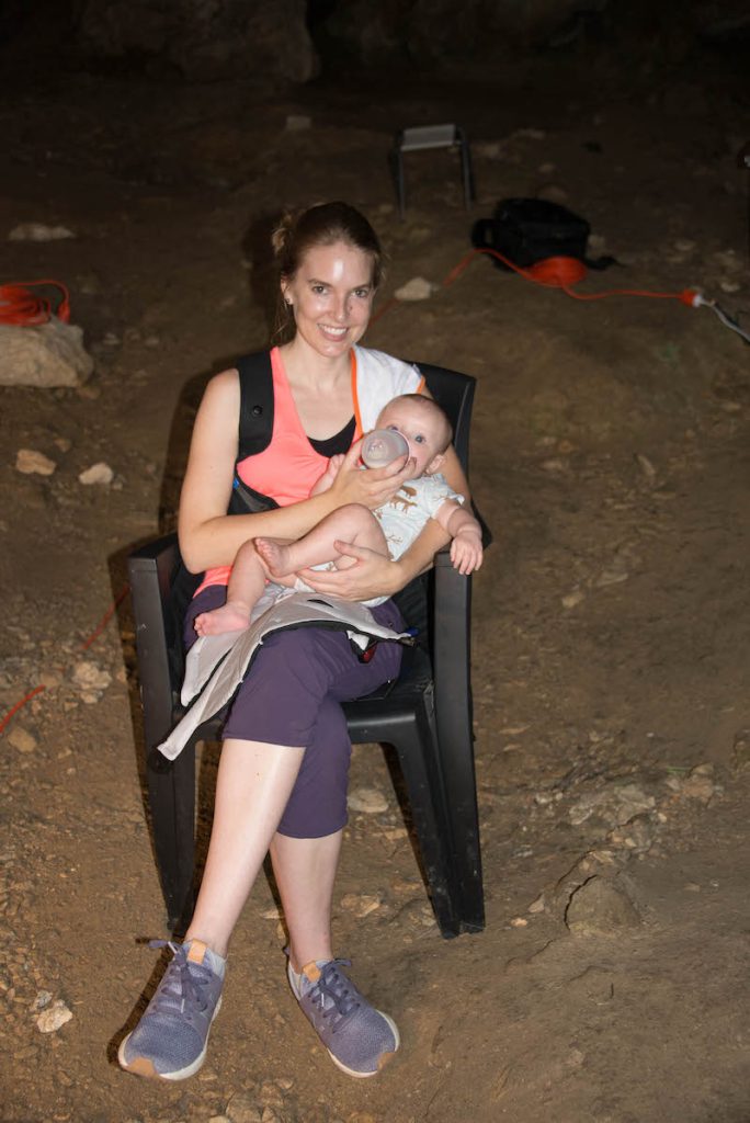 A woman wearing an orange shirt, tights, and running shoes sits on a plastic chair and bottle feeds a baby.