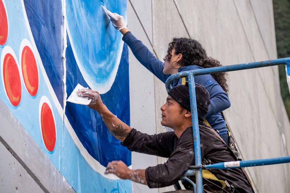 A person with dark curly hair and a person wearing a black hat lean through and over blue metal bars and use white tools to flatten and smooth painted fabric on a concrete wall.