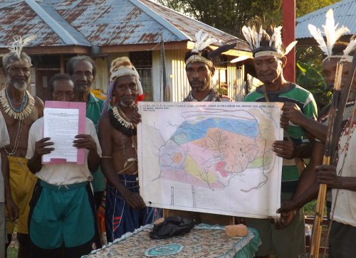 A group of people, some of whom wear white necklaces and white feather headpieces, stand in front of buildings with metal roofs and hold a large, colorful map.