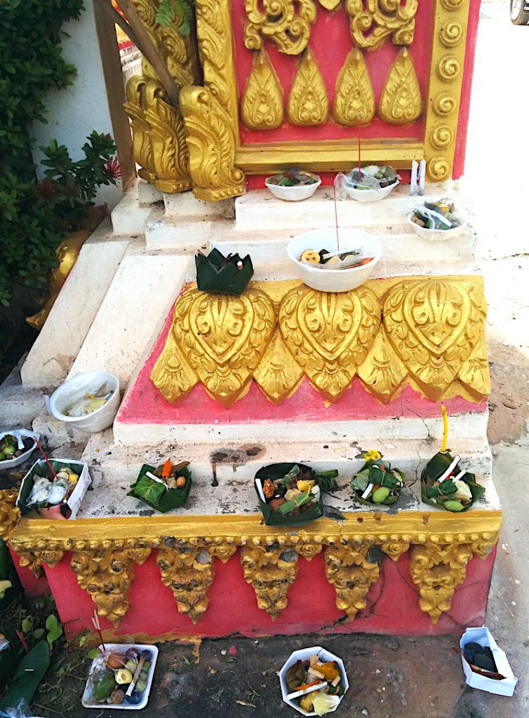 Small containers of food rest on the ground and on the ledges of a pink, white, and gold shrine.