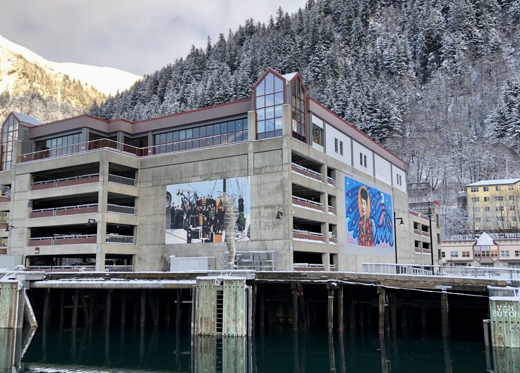 A photo taken from the water shows two murals on two different walls of a building at the base of a hill filled with snow-covered trees.