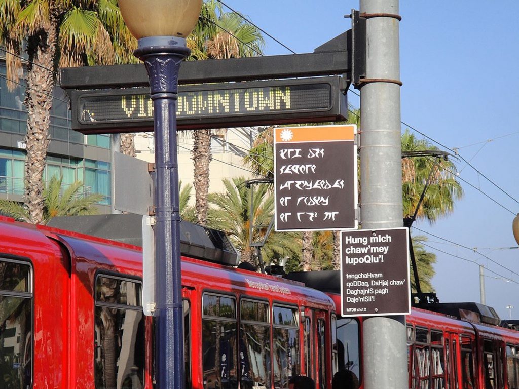 A red bus is parked under a digital street sign that reads “via downtown” and two smaller street signs, each written in a different language.