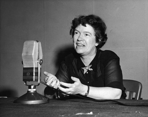 A seated woman with short hair gestures with her hands and speaks into a large rectangular microphone positioned in front of her on a table.