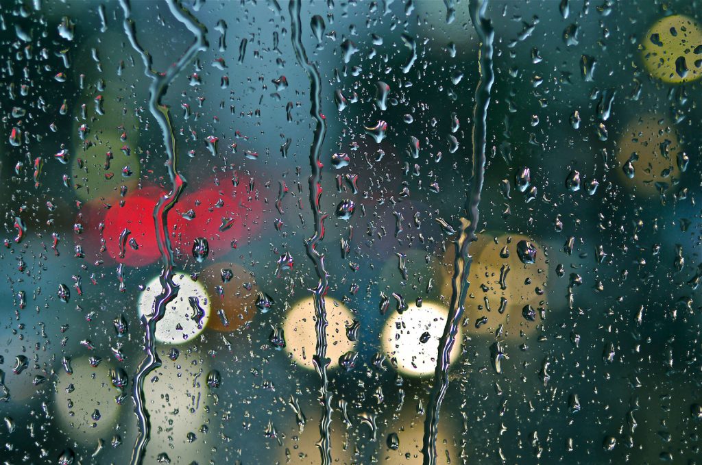 Rain falls down a glass window in front of a blurred background of red and yellow lights.