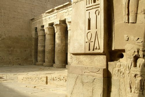 Four large ancient Egyptian hieroglyphs are engraved on the wall of a temple with columns in the background.
