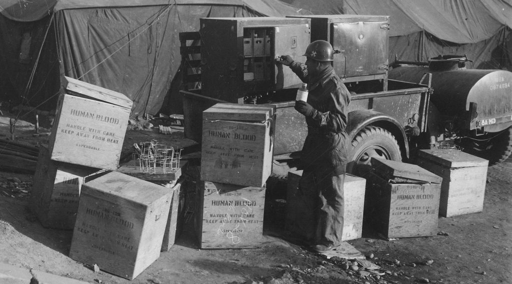 A soldier wearing a helmet and uniform stands in front of a military truck. He is surrounded by several wooden crates labeled “Human blood. Handle with care. Keep away from heat.”