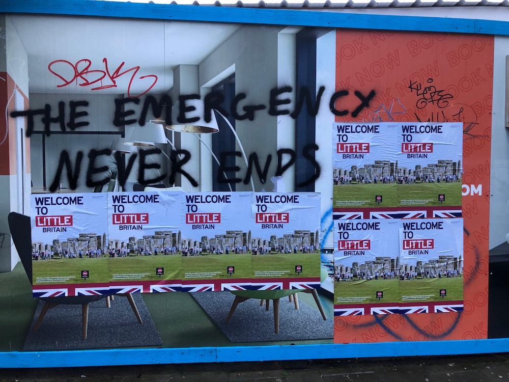 Several copies of the same poster featuring a photograph of Stonehenge and text that reads “Welcome to Little Britain” cover a window. Above them, large black graffiti reads “The Emergency Never Ends.”